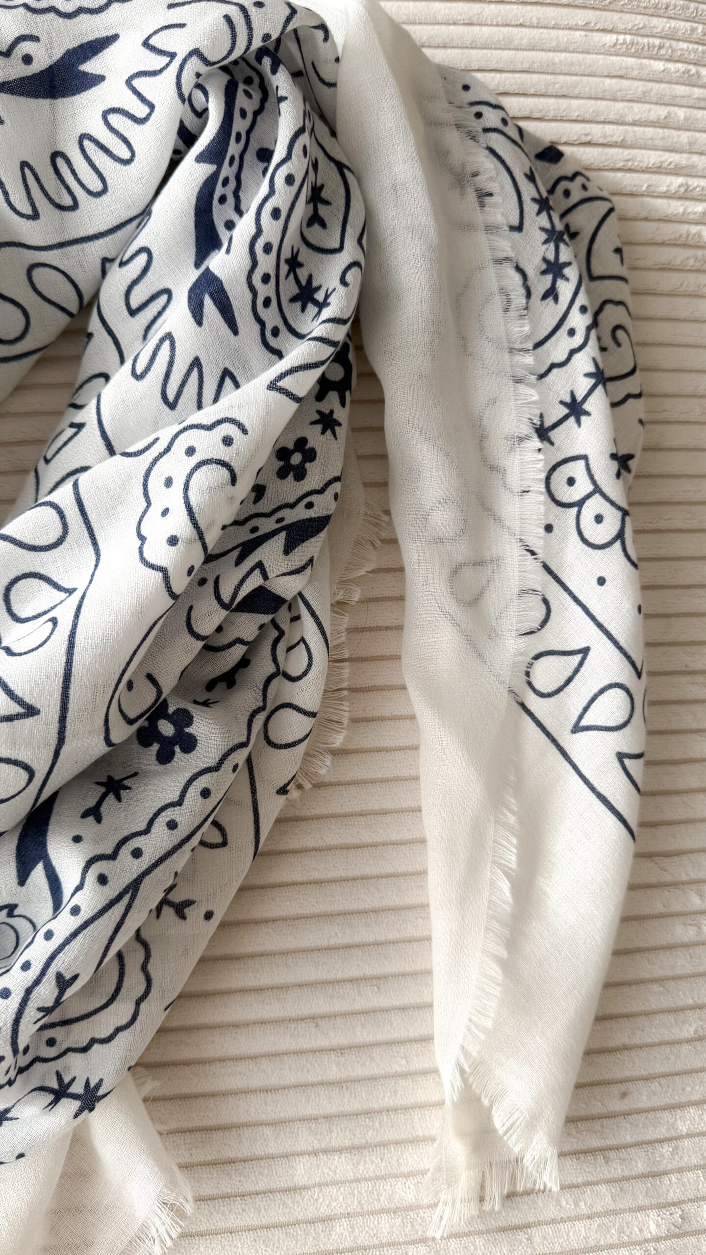 Foulard blanc à motif cachemire noir porté avec un pull crème et un pantalon blanc.