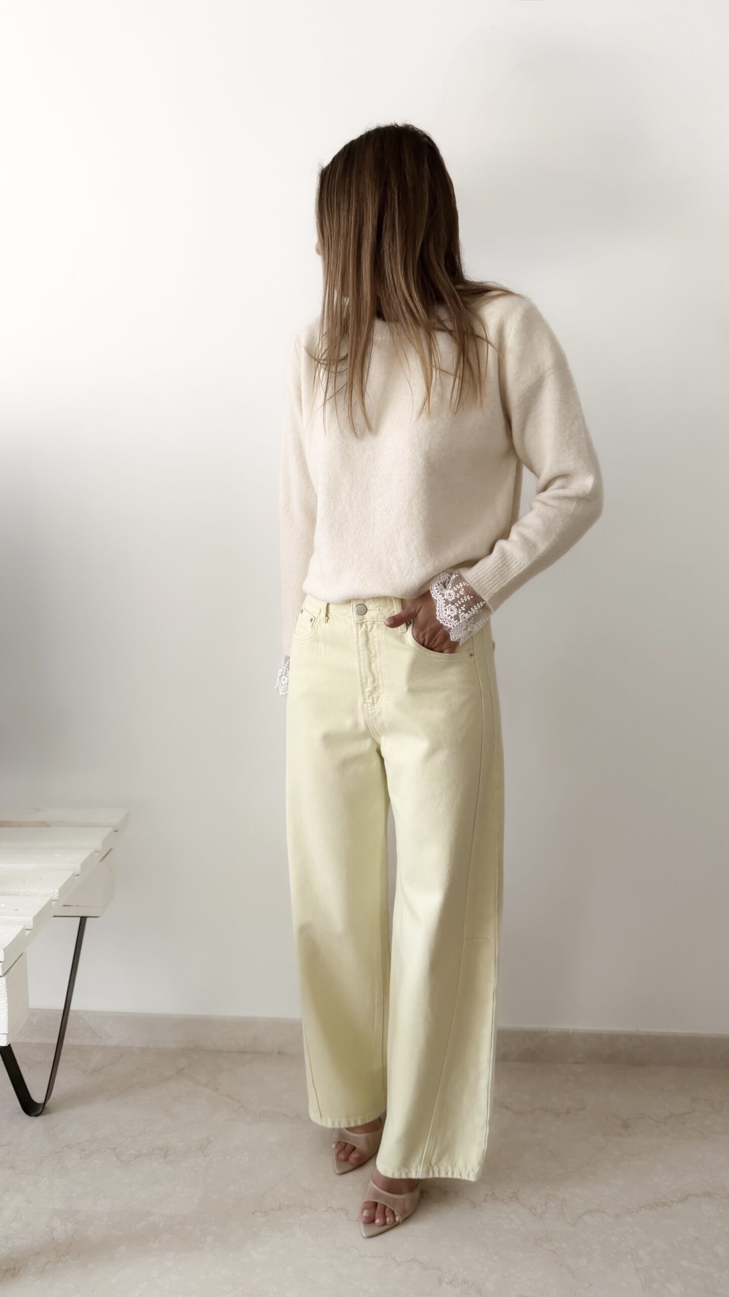 Femme portant un pull crème à détails dentelle aux poignets et un jean large jaune pâle, avec sandales beiges.