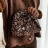 Femme tenant un sac à sequins chocolat scintillant, assorti à une tenue brune élégante avec chemisier et gilet en laine.