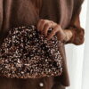 Femme tenant un sac à sequins chocolat scintillant, assorti à une tenue brune élégante avec chemisier et gilet en laine.