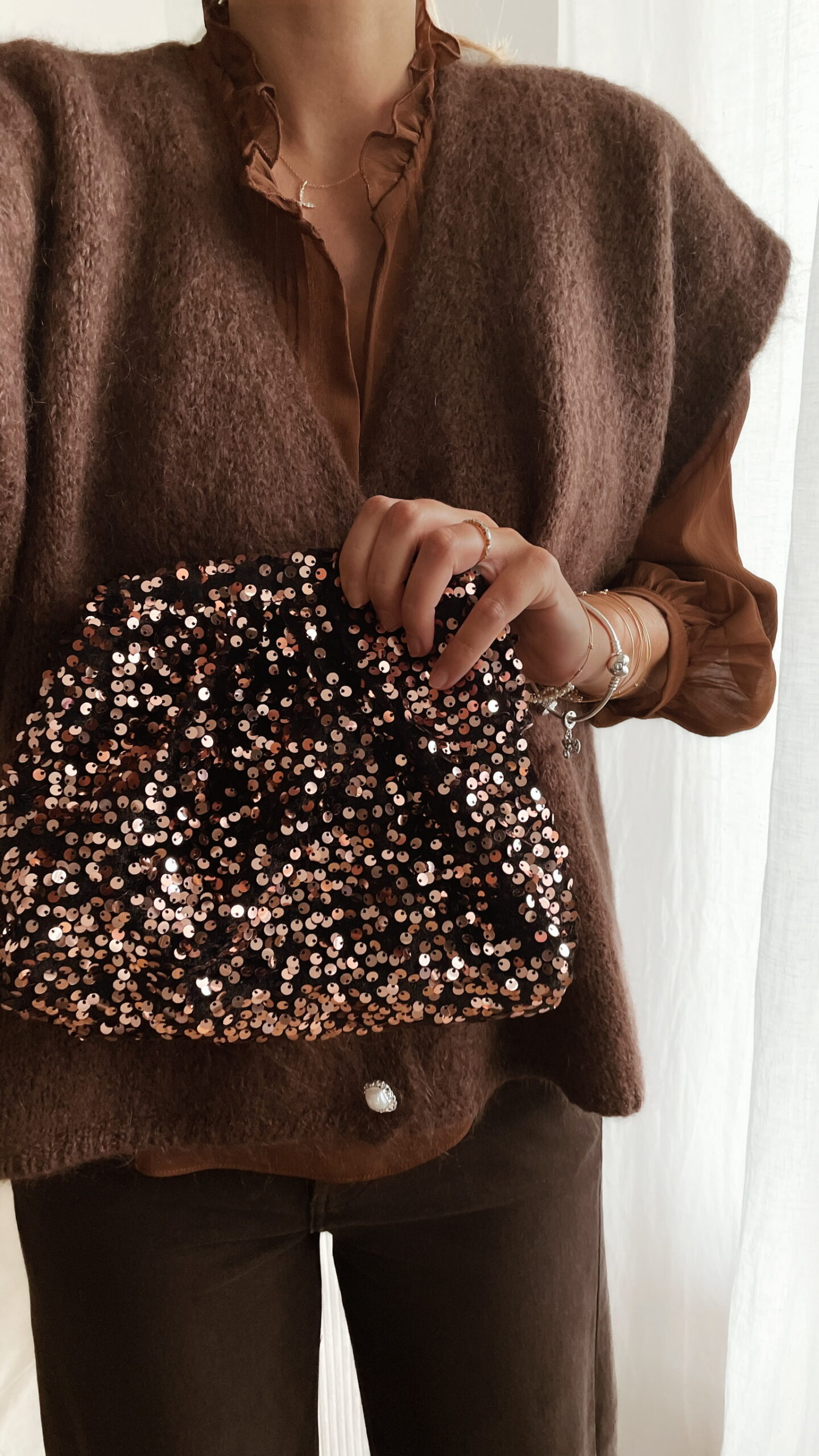 Femme tenant un sac à sequins chocolat scintillant, assorti à une tenue brune élégante avec chemisier et gilet en laine.