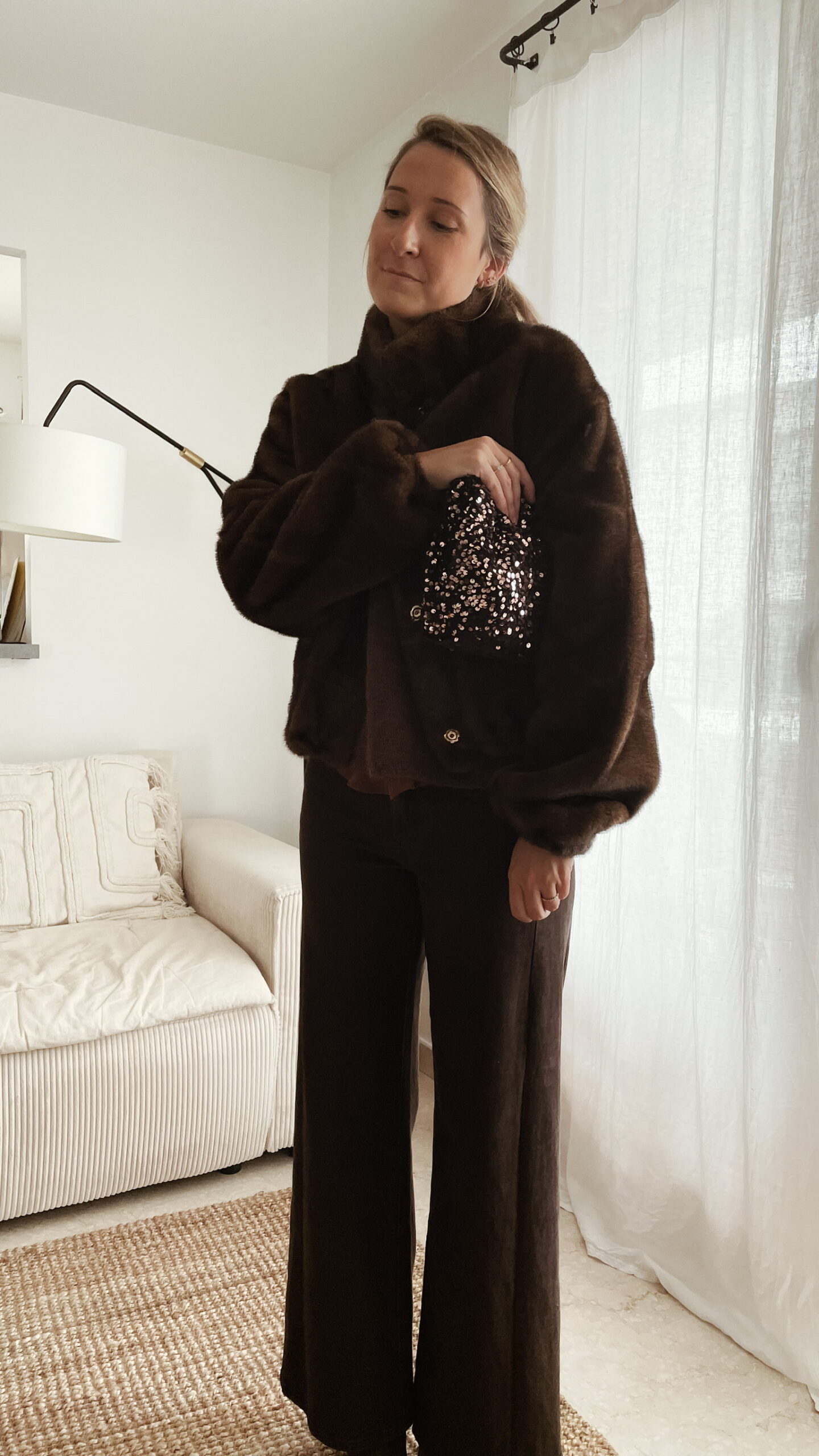 Femme tenant un sac à sequins chocolat scintillant, assorti à une tenue brune élégante avec chemisier et gilet en laine.