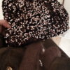 Femme tenant un sac à sequins chocolat scintillant, assorti à une tenue brune élégante avec chemisier et gilet en laine.