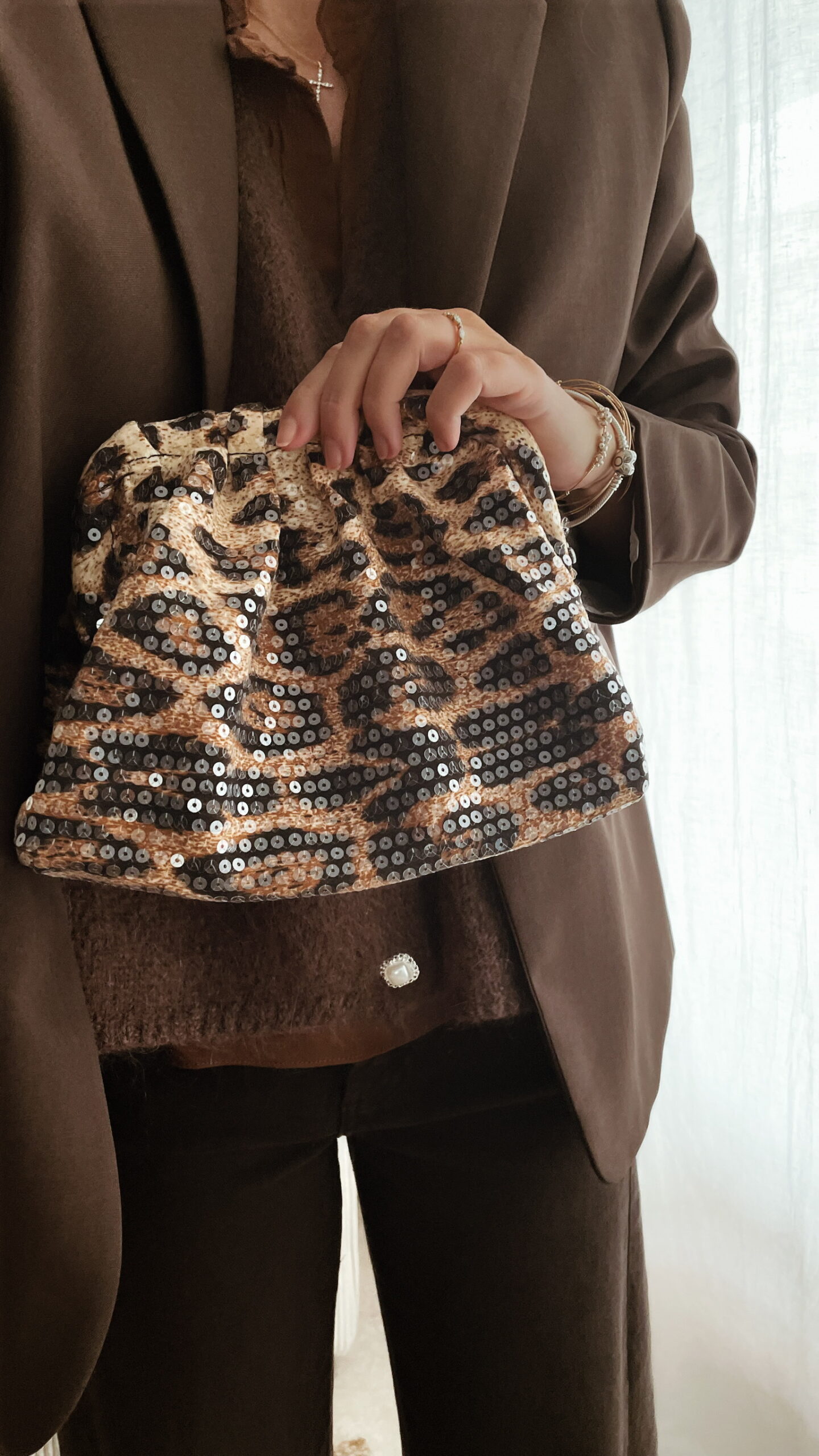 Femme tenant un sac à sequins léopard scintillant, assorti à une tenue brune élégante avec chemisier et blazer en laine.