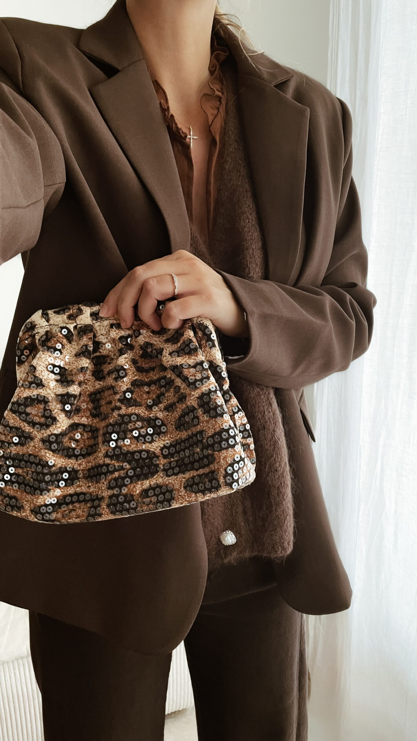 Femme tenant un sac à sequins léopard scintillant, assorti à une tenue brune élégante avec chemisier et blazer en laine.