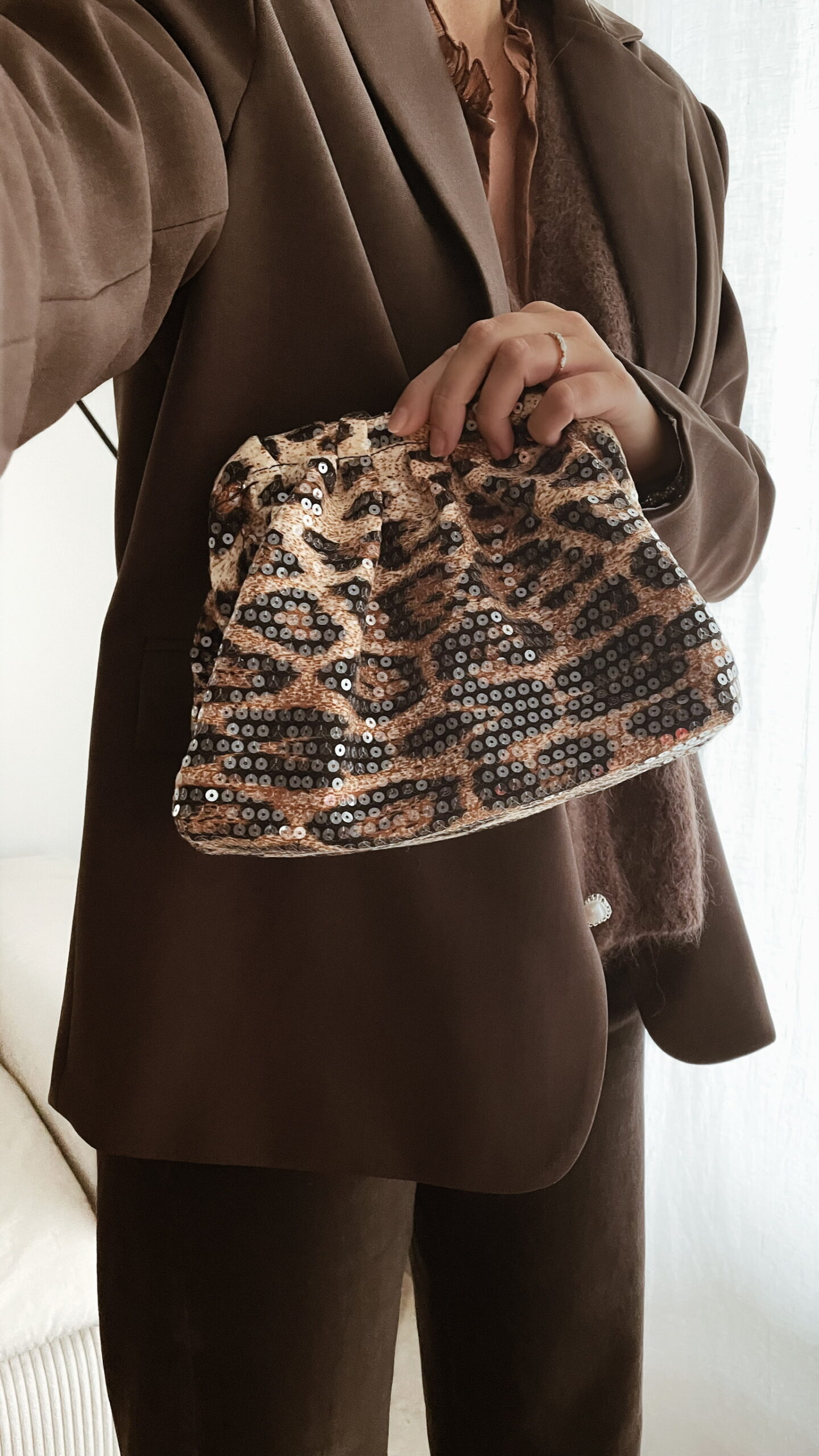 Femme tenant un sac à sequins léopard scintillant, assorti à une tenue brune élégante avec chemisier et blazer en laine.