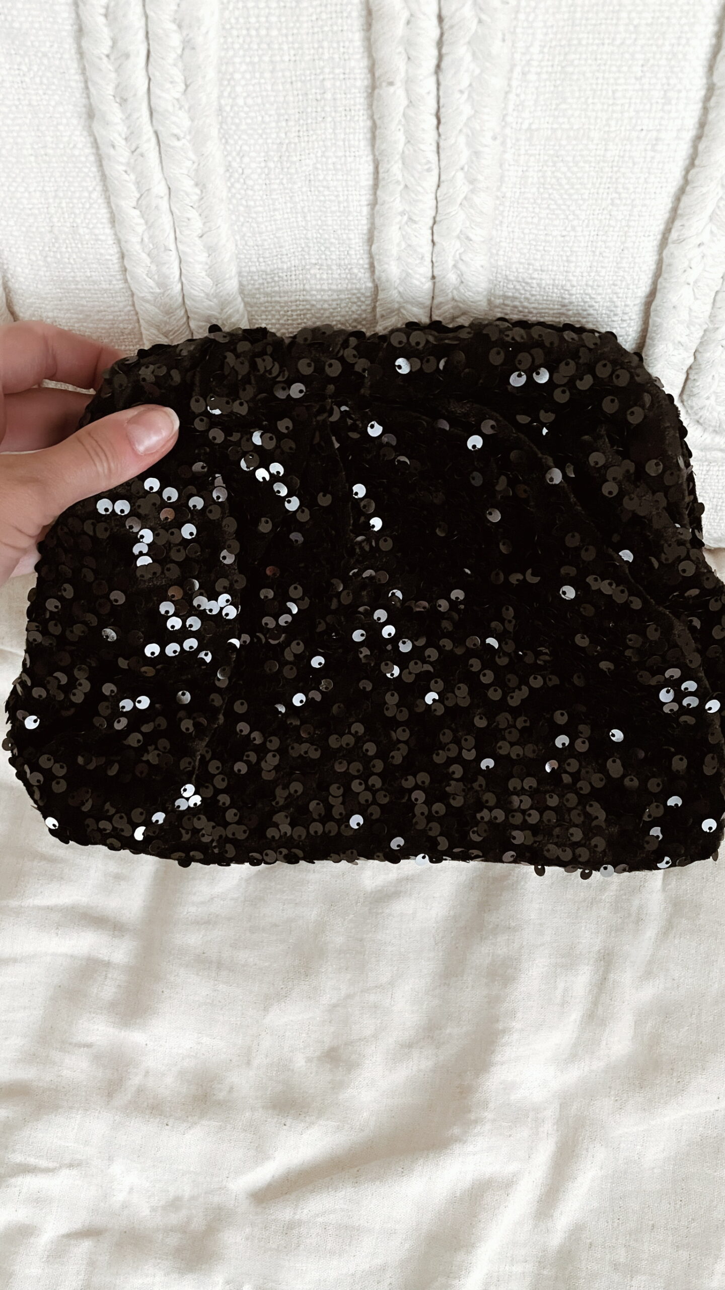 Femme tenant un sac à sequins noir scintillant, assorti à une tenue brune élégante avec chemisier et blazer en laine.