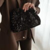 Femme tenant un sac à sequins noir scintillant, assorti à une tenue brune élégante avec chemisier et blazer en laine.