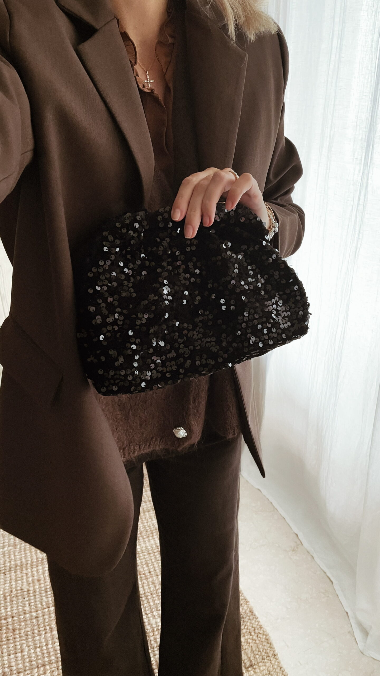 Femme tenant un sac à sequins noir scintillant, assorti à une tenue brune élégante avec chemisier et blazer en laine.