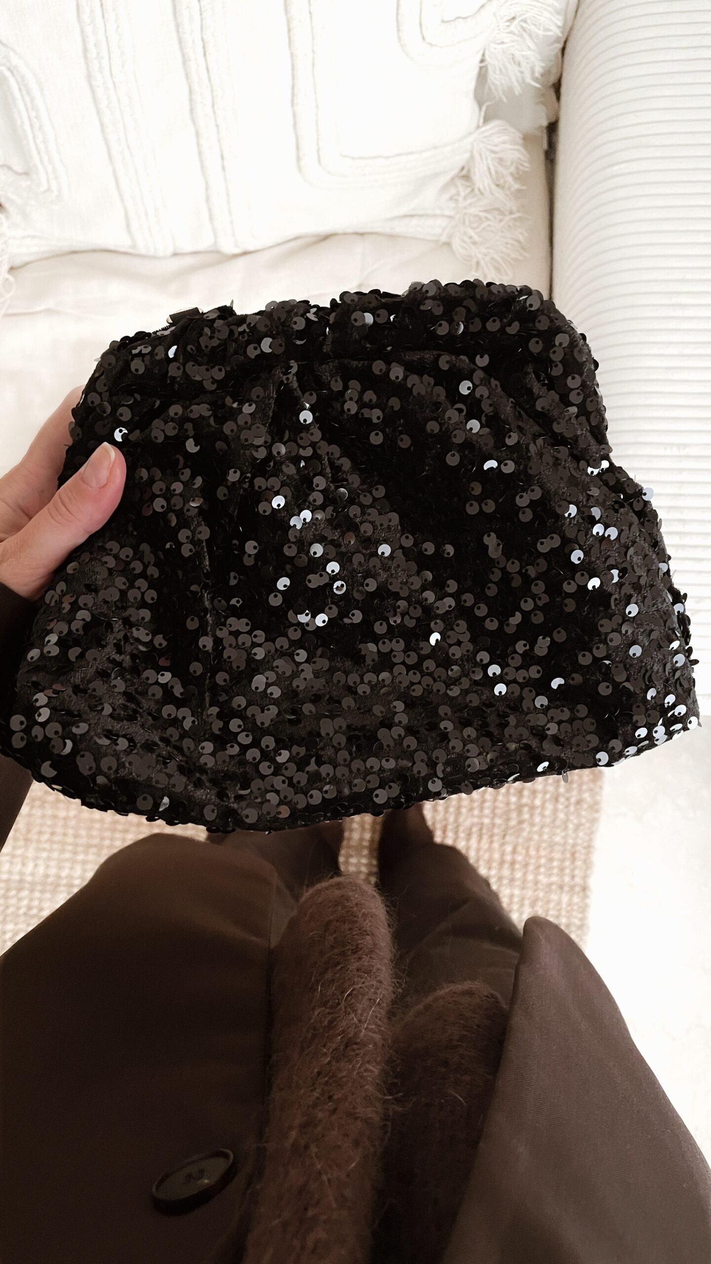 Femme tenant un sac à sequins noir scintillant, assorti à une tenue brune élégante avec chemisier et blazer en laine.