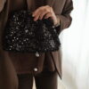 Femme tenant un sac à sequins noir scintillant, assorti à une tenue brune élégante avec chemisier et blazer en laine.