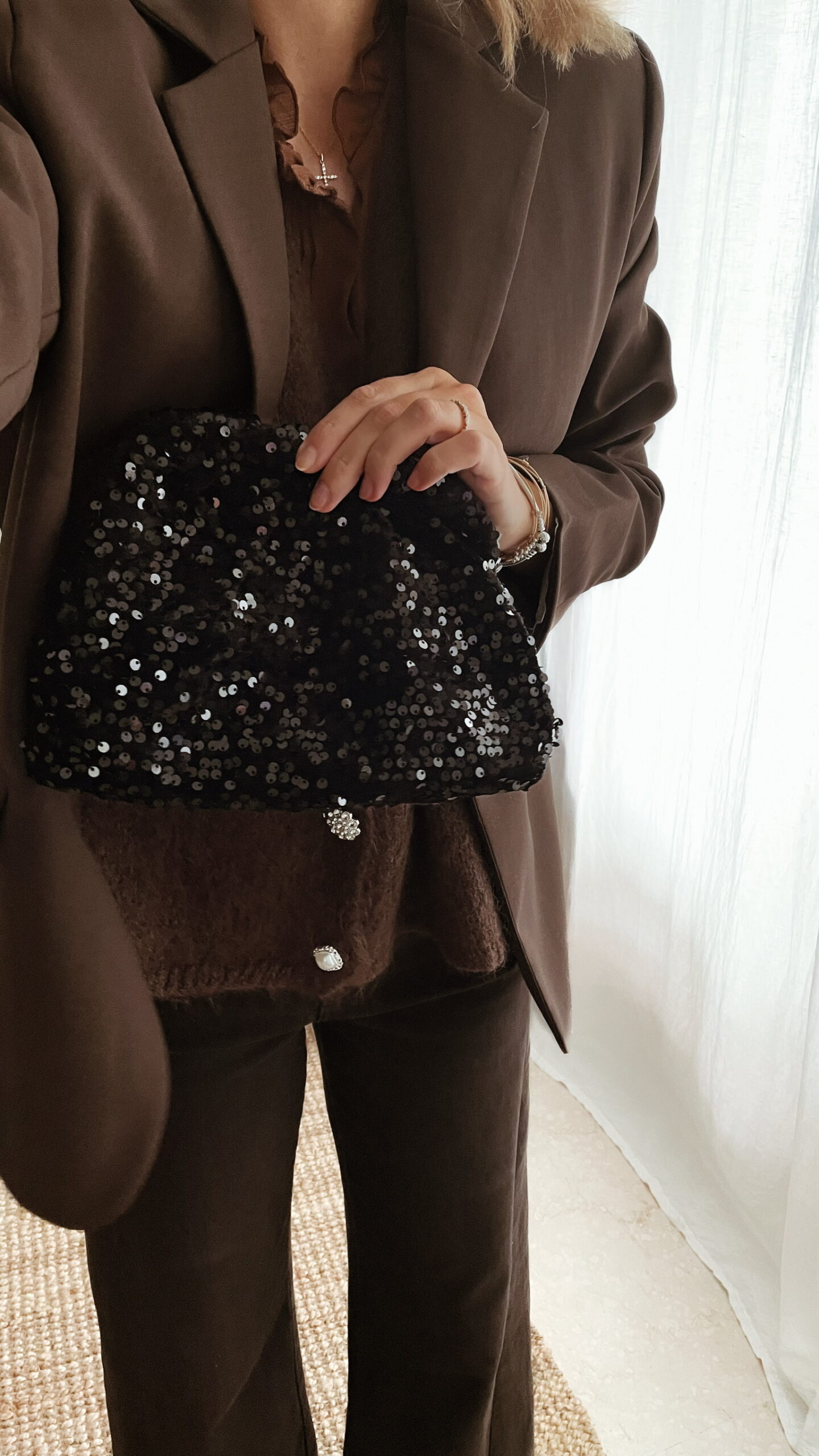 Femme tenant un sac à sequins noir scintillant, assorti à une tenue brune élégante avec chemisier et blazer en laine.
