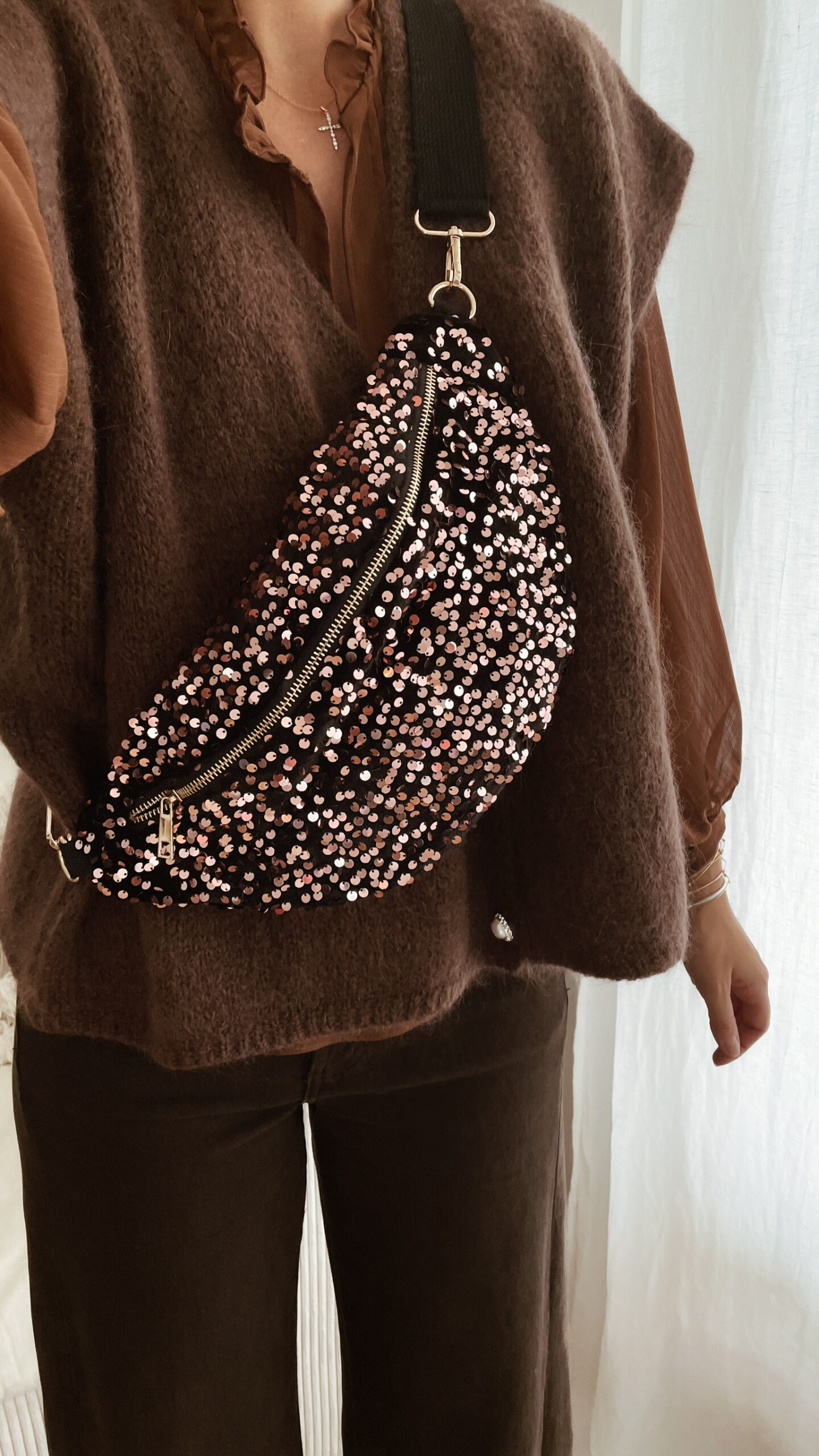 Femme tenant un sac banane à sequins chocolat scintillant, assorti à une tenue brune élégante avec chemisier et gilet en laine.
