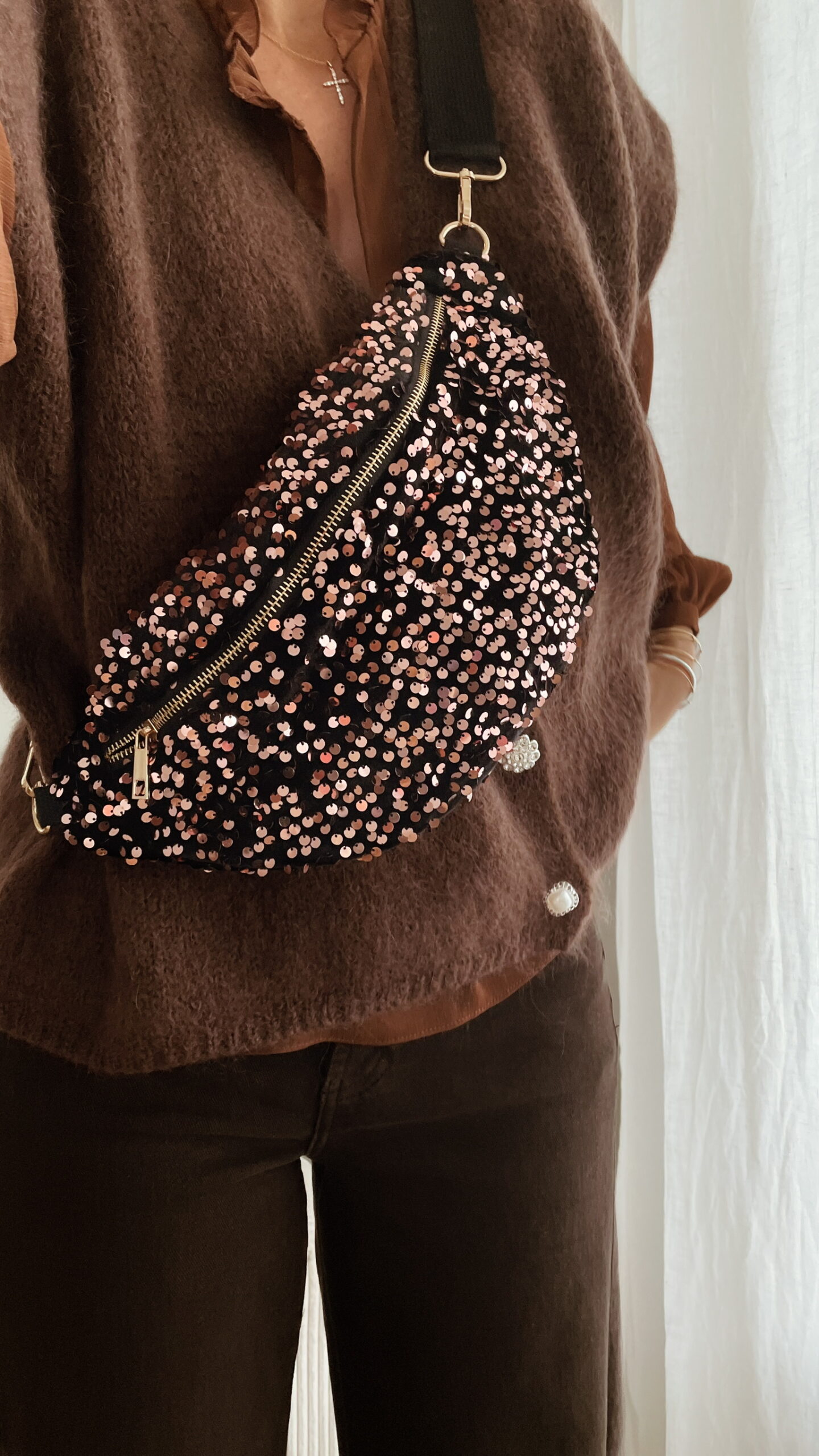 Femme tenant un sac banane à sequins chocolat scintillant, assorti à une tenue brune élégante avec chemisier et gilet en laine.