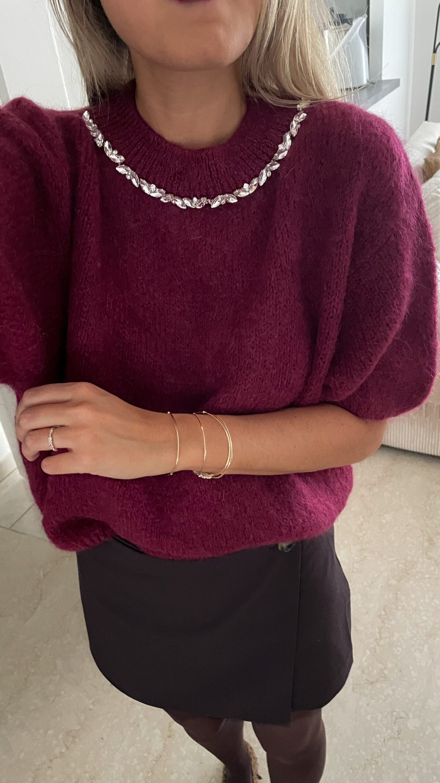 Femme portant un pull en maille bordeaux orné de strass au col, associé à une jupe noire courte et des bijoux dorés.
