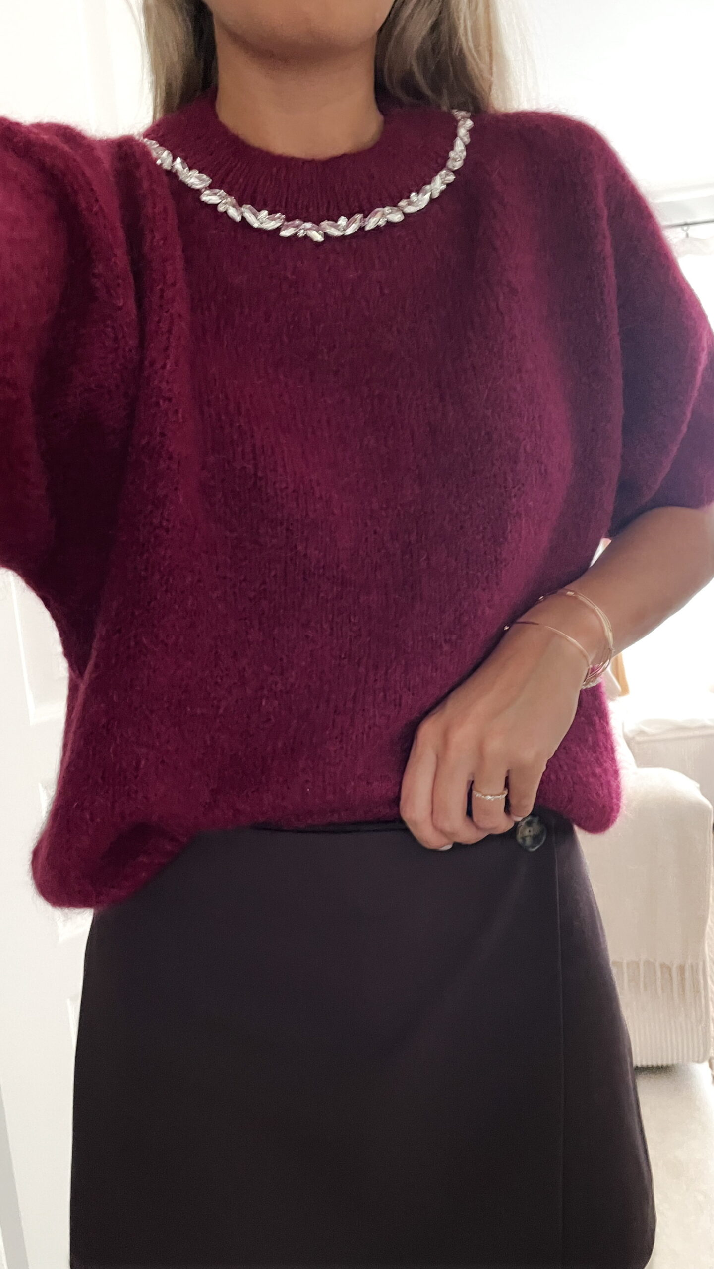 Femme portant un pull en maille bordeaux orné de strass au col, associé à une jupe noire courte et des bijoux dorés.