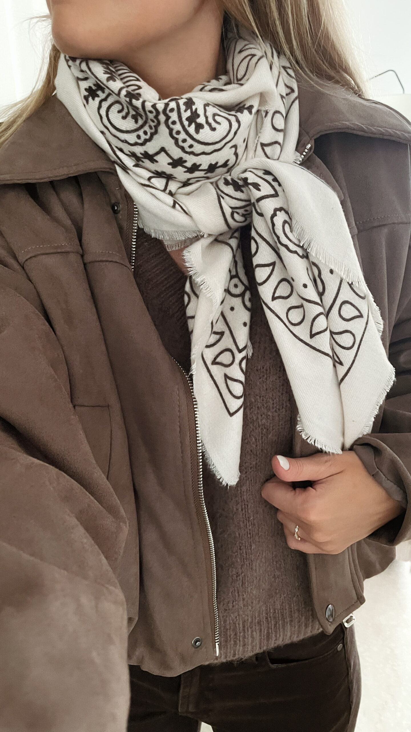Femme portant une veste effet daim couleur marron taupe, assortie à un pull en alpaga taupe ton sur ton, un pantalon marron, une echarpe à motifs et un sac en daim marron avec des franges dans un intérieur lumineux.