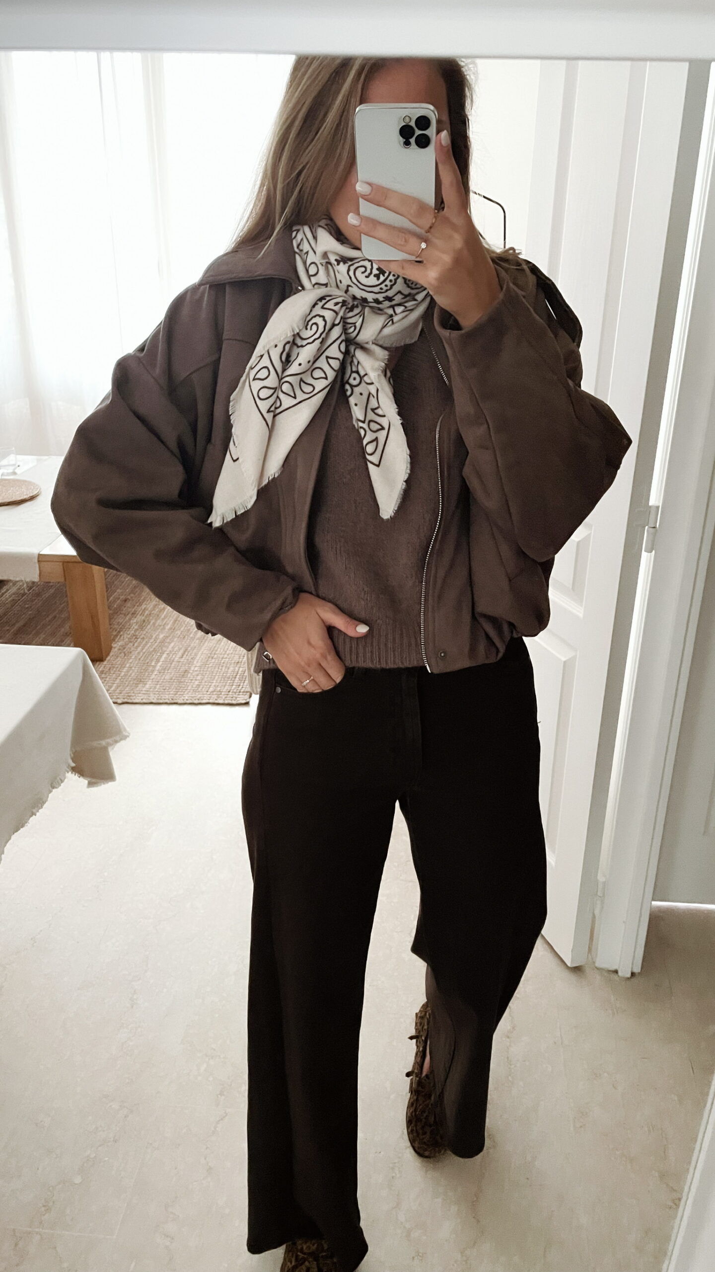 Femme portant une veste effet daim couleur marron taupe, assortie à un pull en alpaga taupe ton sur ton, un pantalon marron, une echarpe à motifs et un sac en daim marron avec des franges dans un intérieur lumineux.