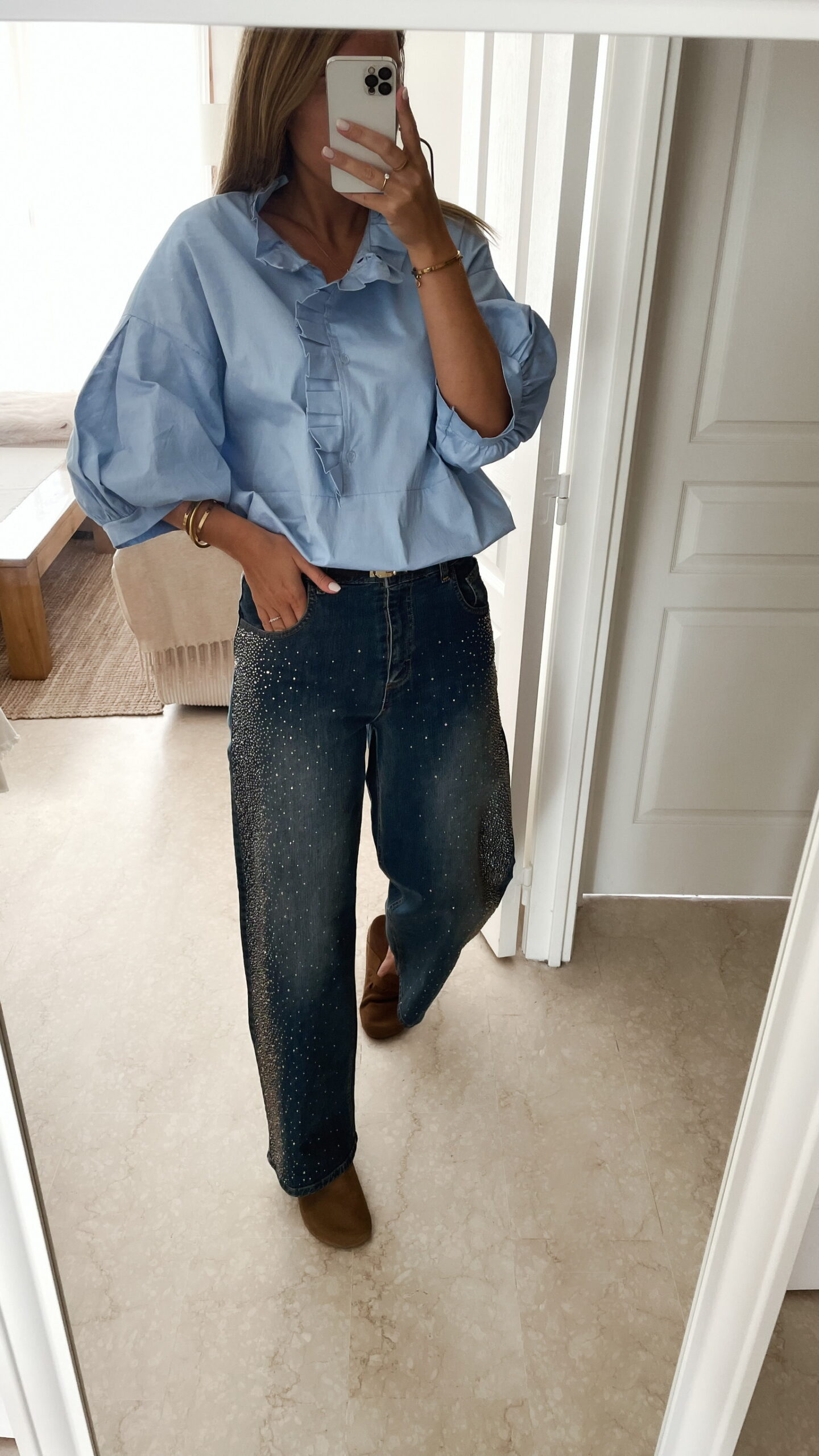 Femme portant une chemise oversize bleu clair à volants et un jean bleu orné de strass sur les côtés, dans un intérieur lumineux et minimaliste.