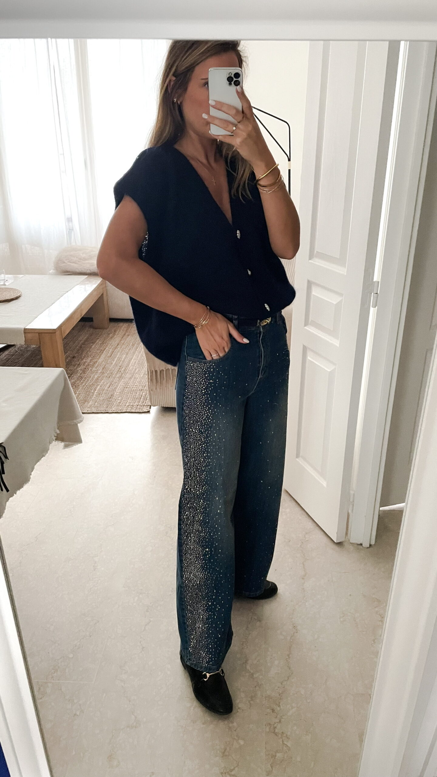 Femme portant une chemise oversize bleu clair à volants et un jean bleu orné de strass sur les côtés, dans un intérieur lumineux et minimaliste.