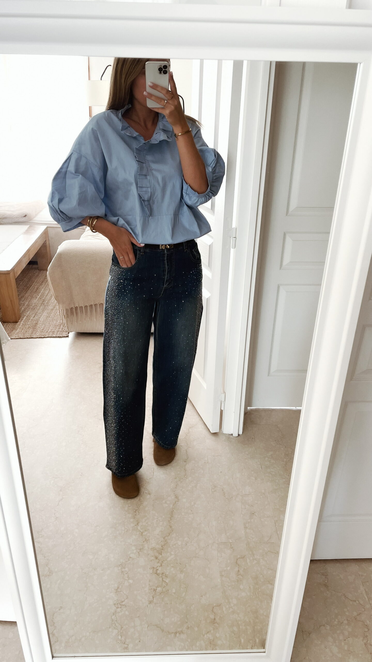Femme portant une chemise oversize bleu clair à volants et un jean bleu orné de strass sur les côtés, dans un intérieur lumineux et minimaliste.