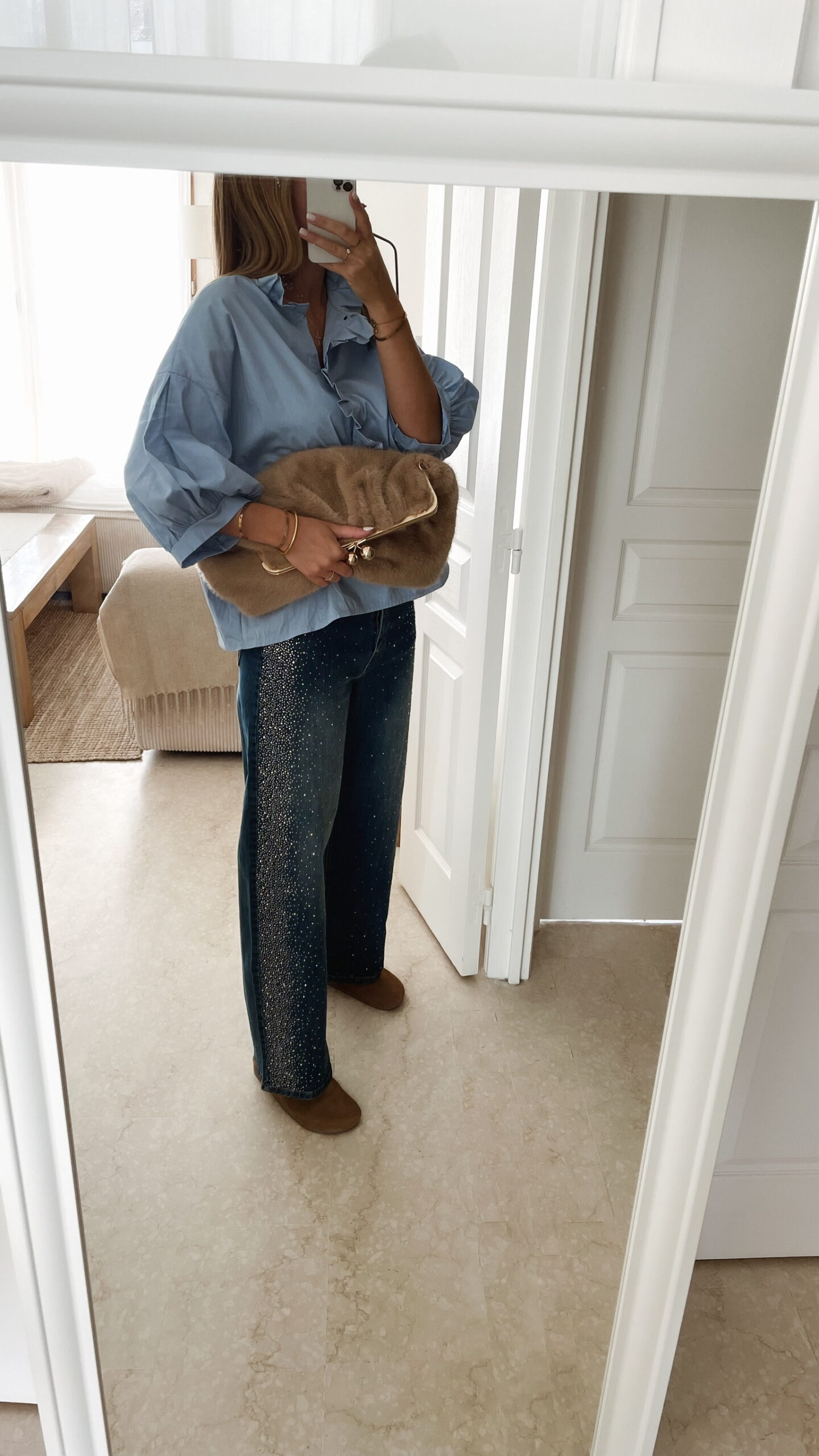 Femme portant une chemise oversize bleu clair à volants et un jean bleu orné de strass sur les côtés, dans un intérieur lumineux et minimaliste.