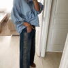 Femme portant une chemise oversize bleu clair à volants et un jean bleu orné de strass sur les côtés, dans un intérieur lumineux et minimaliste.
