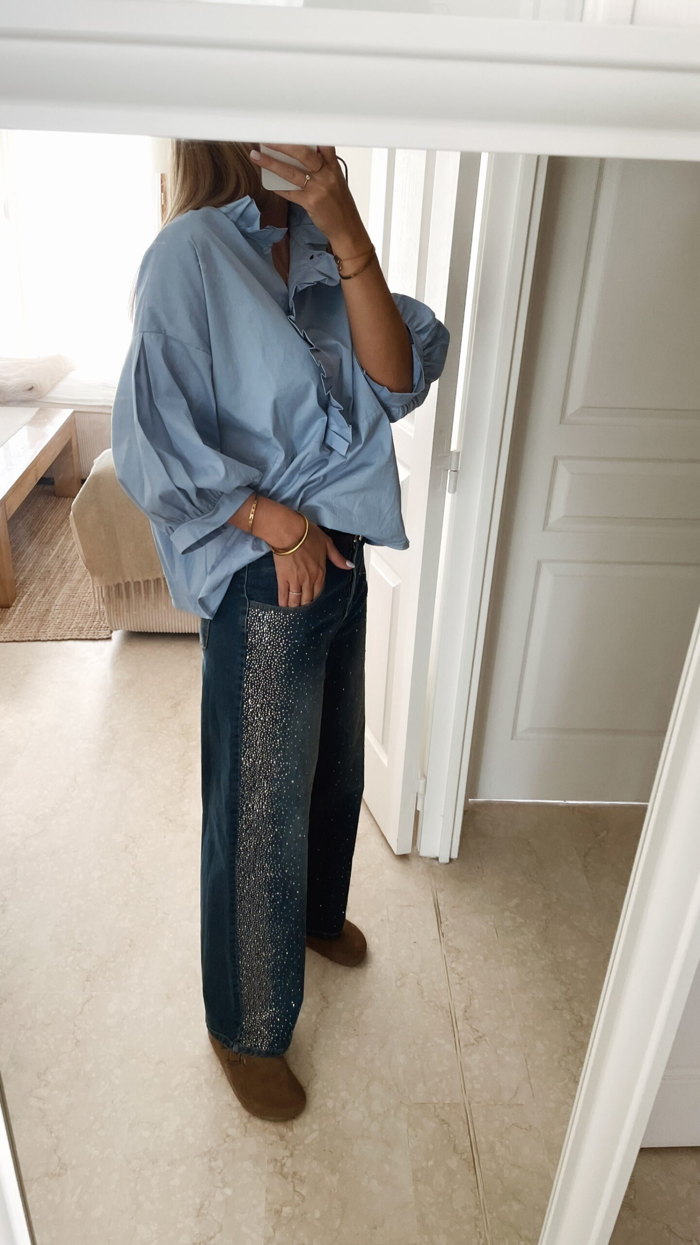 Femme portant une chemise oversize bleu clair à volants et un jean bleu orné de strass sur les côtés, dans un intérieur lumineux et minimaliste.