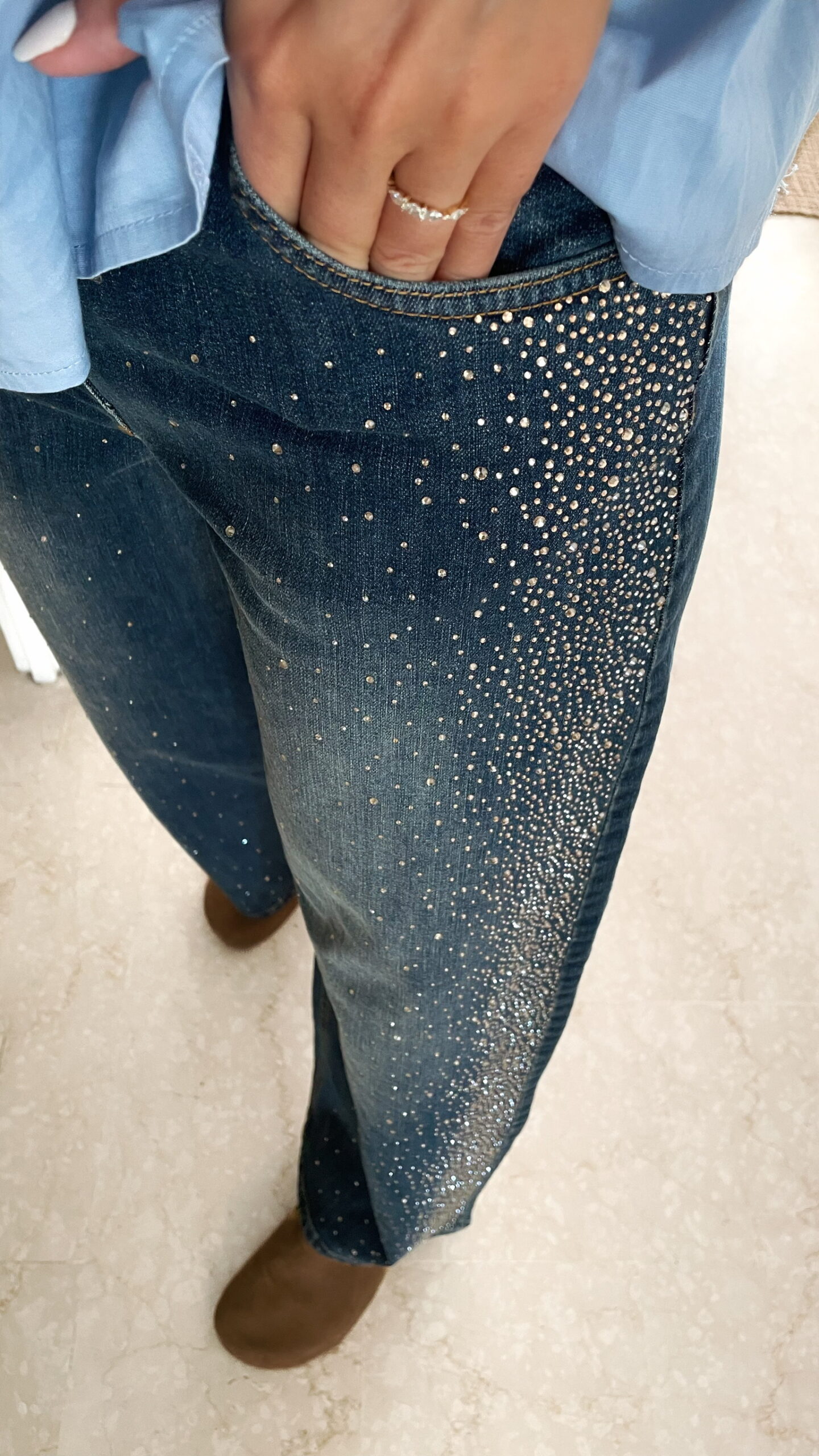 Femme portant une chemise oversize bleu clair à volants et un jean bleu orné de strass sur les côtés, dans un intérieur lumineux et minimaliste.