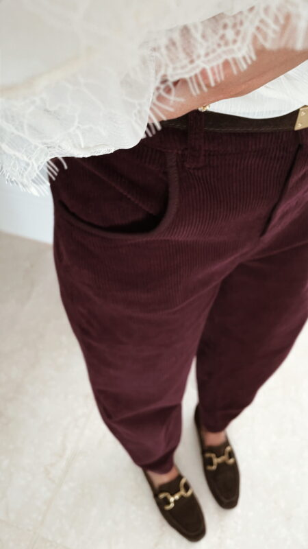 Femme portant un pantalon ballon en velours bordeaux, une blouse blanche à dentelle et un foulard imprimé beige, complété par des mocassins marron à boucle dorée.