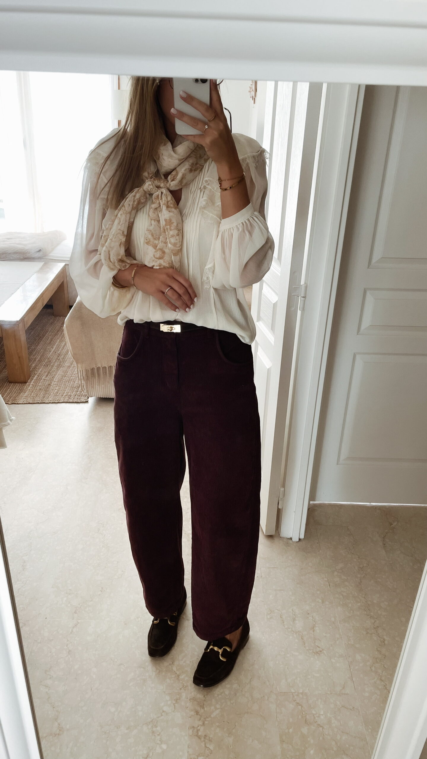Femme portant un pantalon ballon en velours bordeaux, une blouse blanche à dentelle et un foulard imprimé beige, complété par des mocassins marron à boucle dorée.