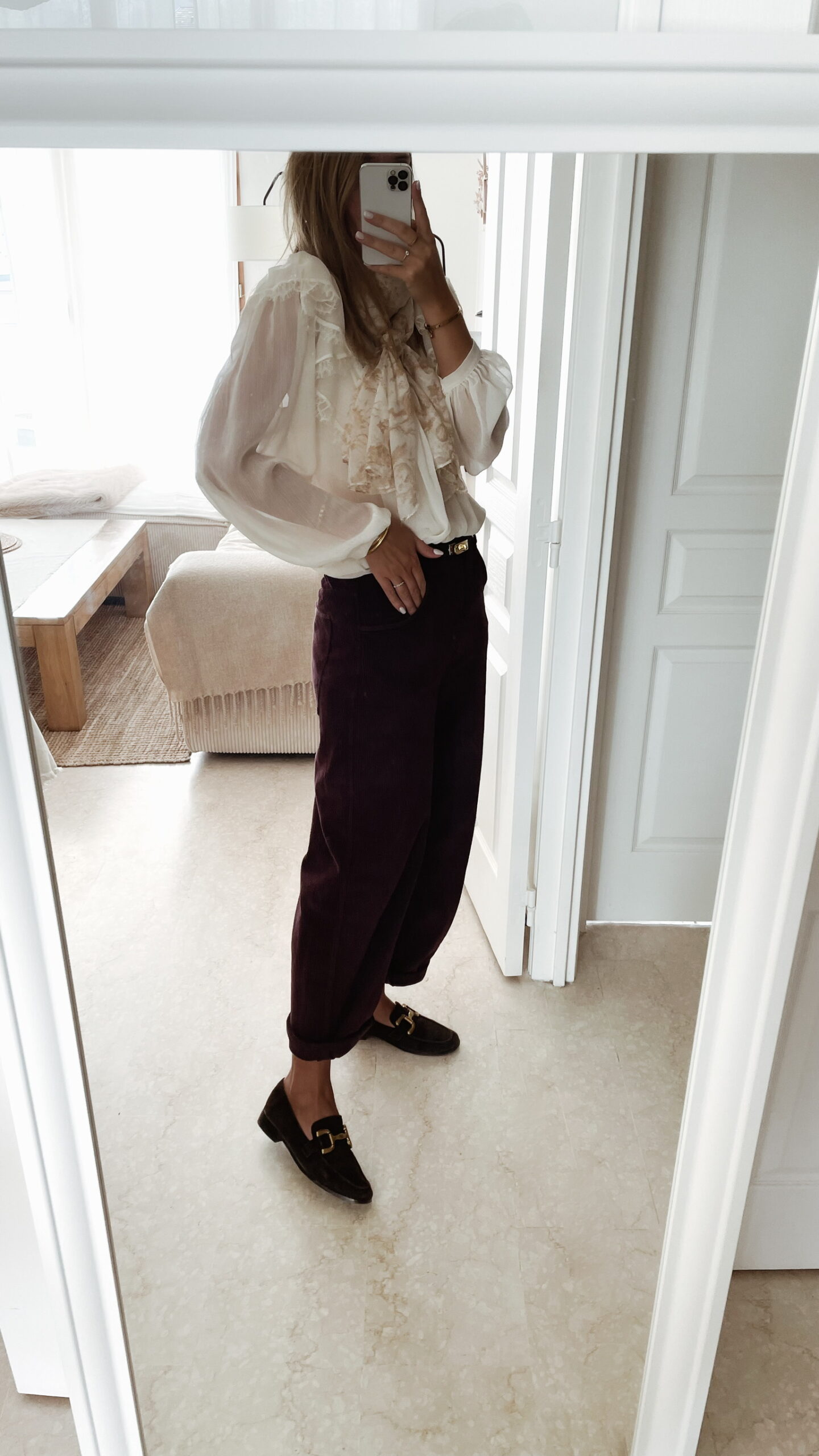 Femme portant un pantalon ballon en velours bordeaux, une blouse blanche à dentelle et un foulard imprimé beige, complété par des mocassins marron à boucle dorée.