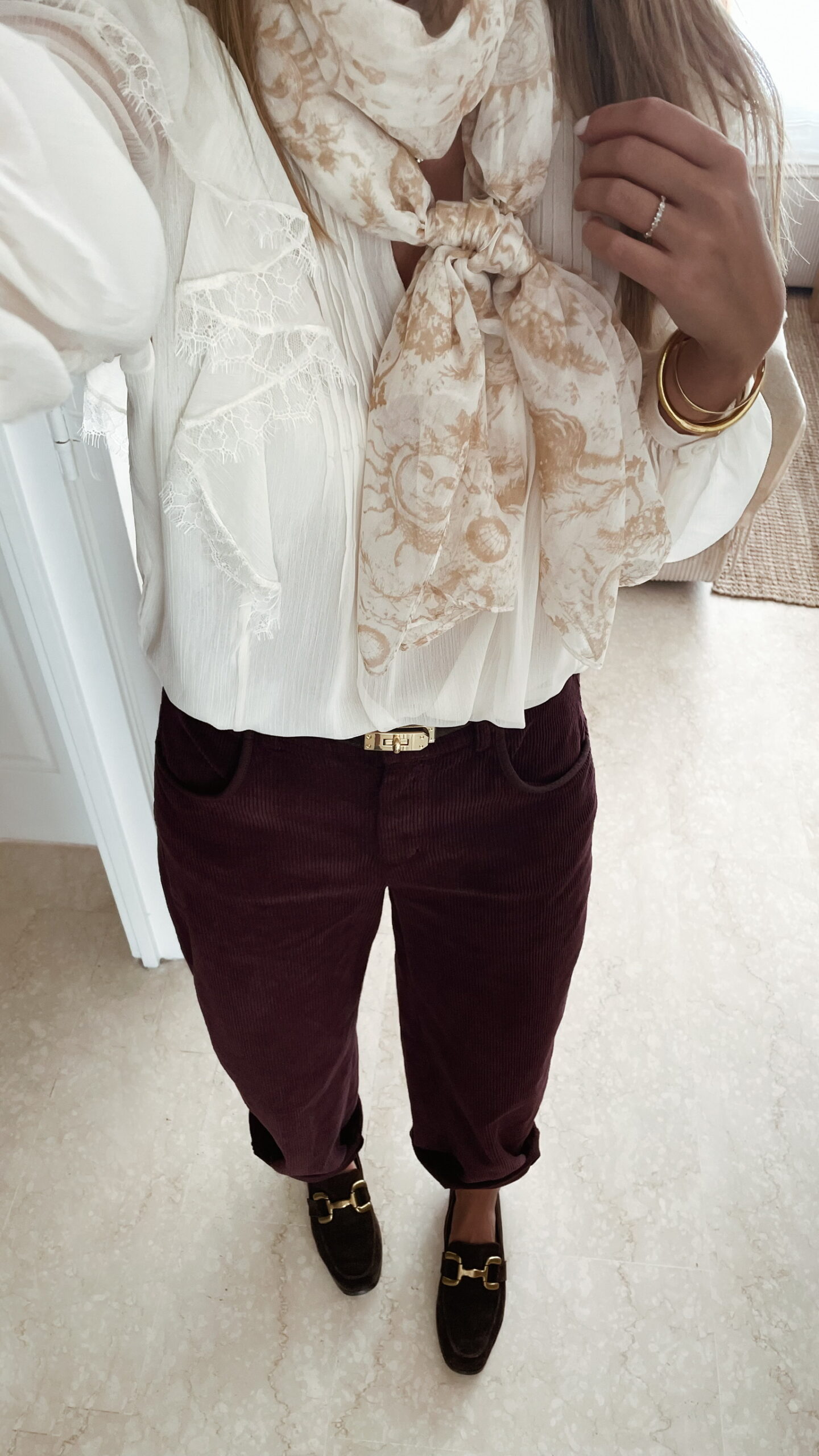 Femme portant un pantalon ballon en velours bordeaux, une blouse blanche à dentelle et un foulard imprimé beige, complété par des mocassins marron à boucle dorée.