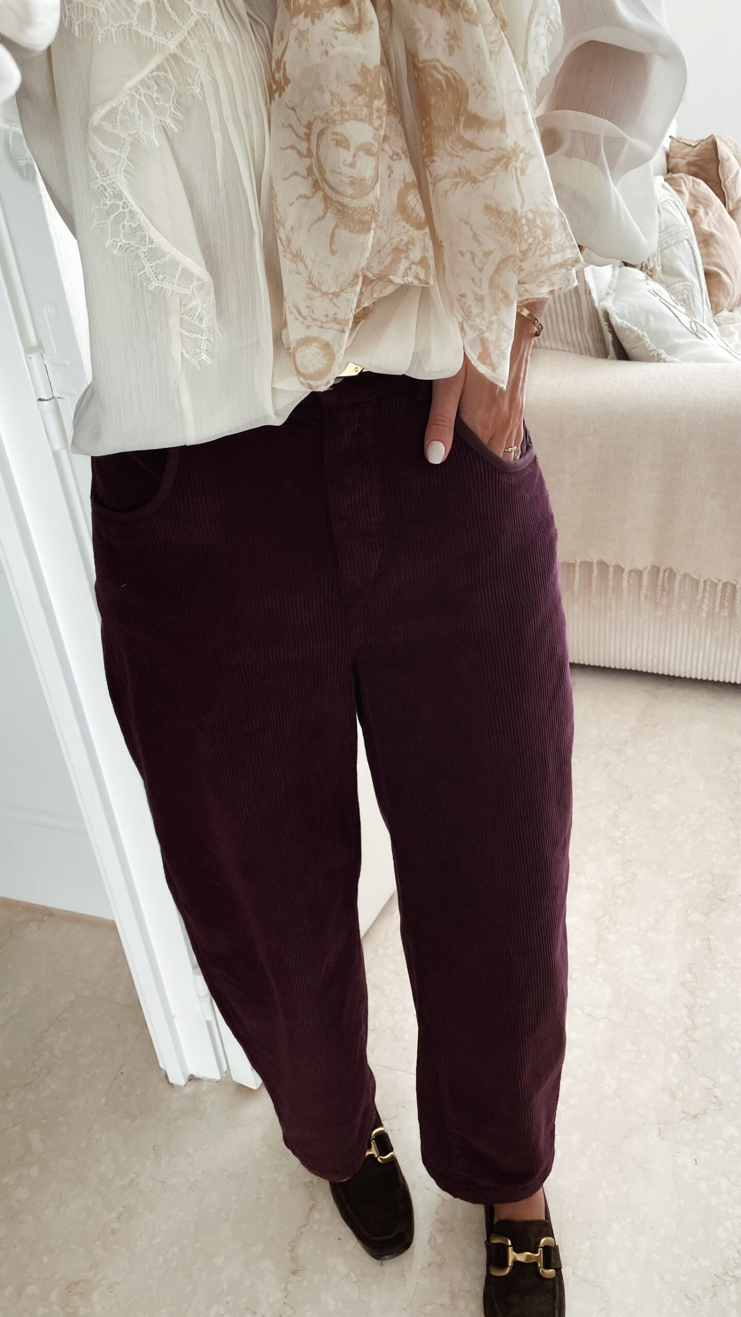 Femme portant un pantalon ballon en velours bordeaux, une blouse blanche à dentelle et un foulard imprimé beige, complété par des mocassins marron à boucle dorée.