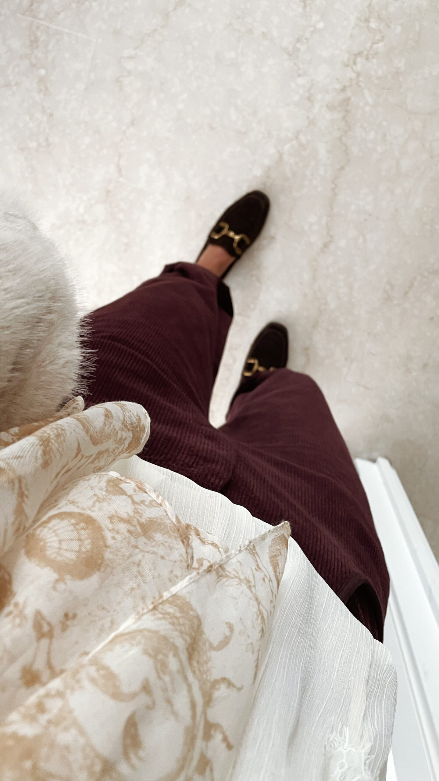 Femme portant un pantalon ballon en velours bordeaux, une blouse blanche à dentelle et un foulard imprimé beige, complété par des mocassins marron à boucle dorée.