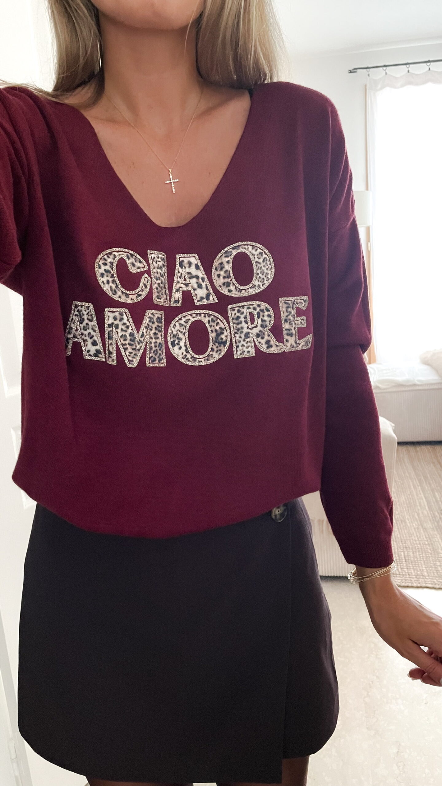 Femme portant un pull bordeaux à inscription “Ciao Amore”, associé à une jupe noire, une écharpe imprimée beige, un sac à franges et des mocassins léopard pour un look automnal chic et décontracté.