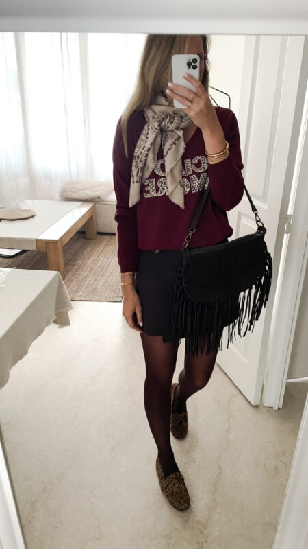Femme portant un pull bordeaux à inscription “Ciao Amore”, associé à une jupe noire, une écharpe imprimée beige, un sac à franges et des mocassins léopard pour un look automnal chic et décontracté.