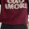 Femme portant un pull bordeaux à inscription “Ciao Amore”, associé à une jupe noire, une écharpe imprimée beige, un sac à franges et des mocassins léopard pour un look automnal chic et décontracté.