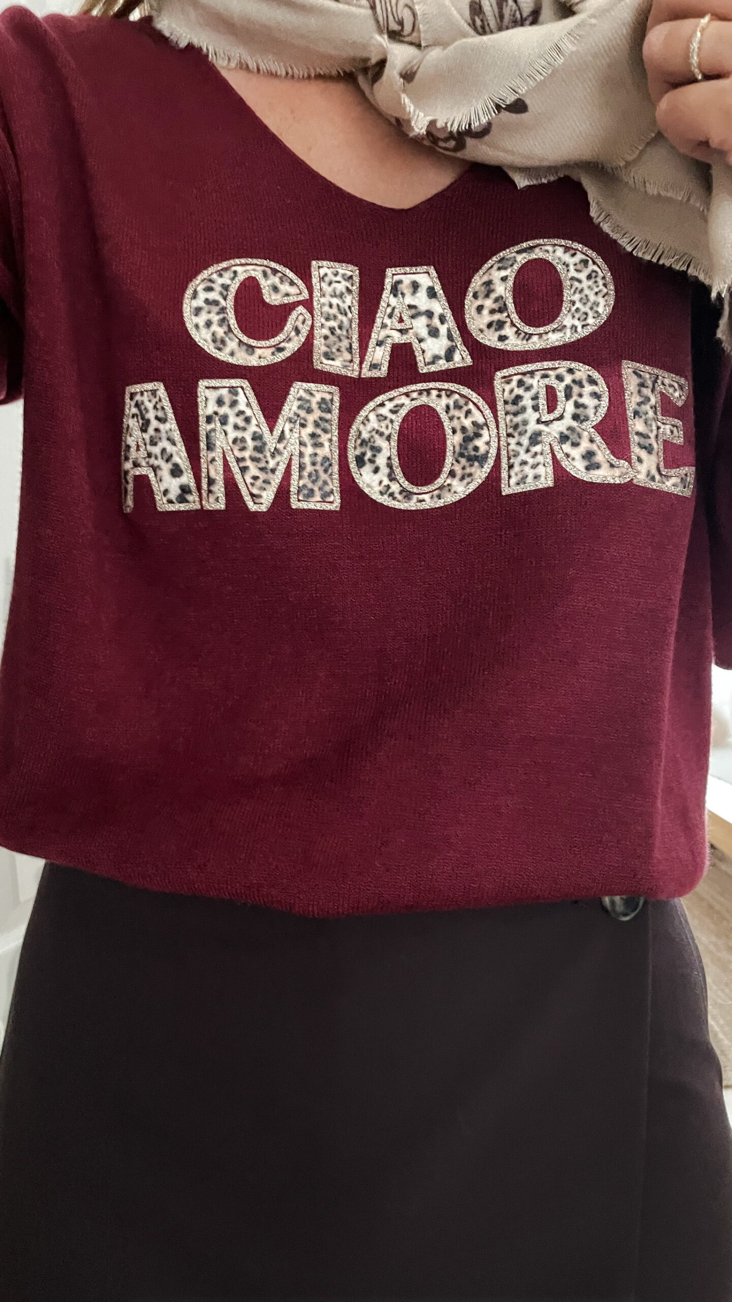Femme portant un pull bordeaux à inscription “Ciao Amore”, associé à une jupe noire, une écharpe imprimée beige, un sac à franges et des mocassins léopard pour un look automnal chic et décontracté.