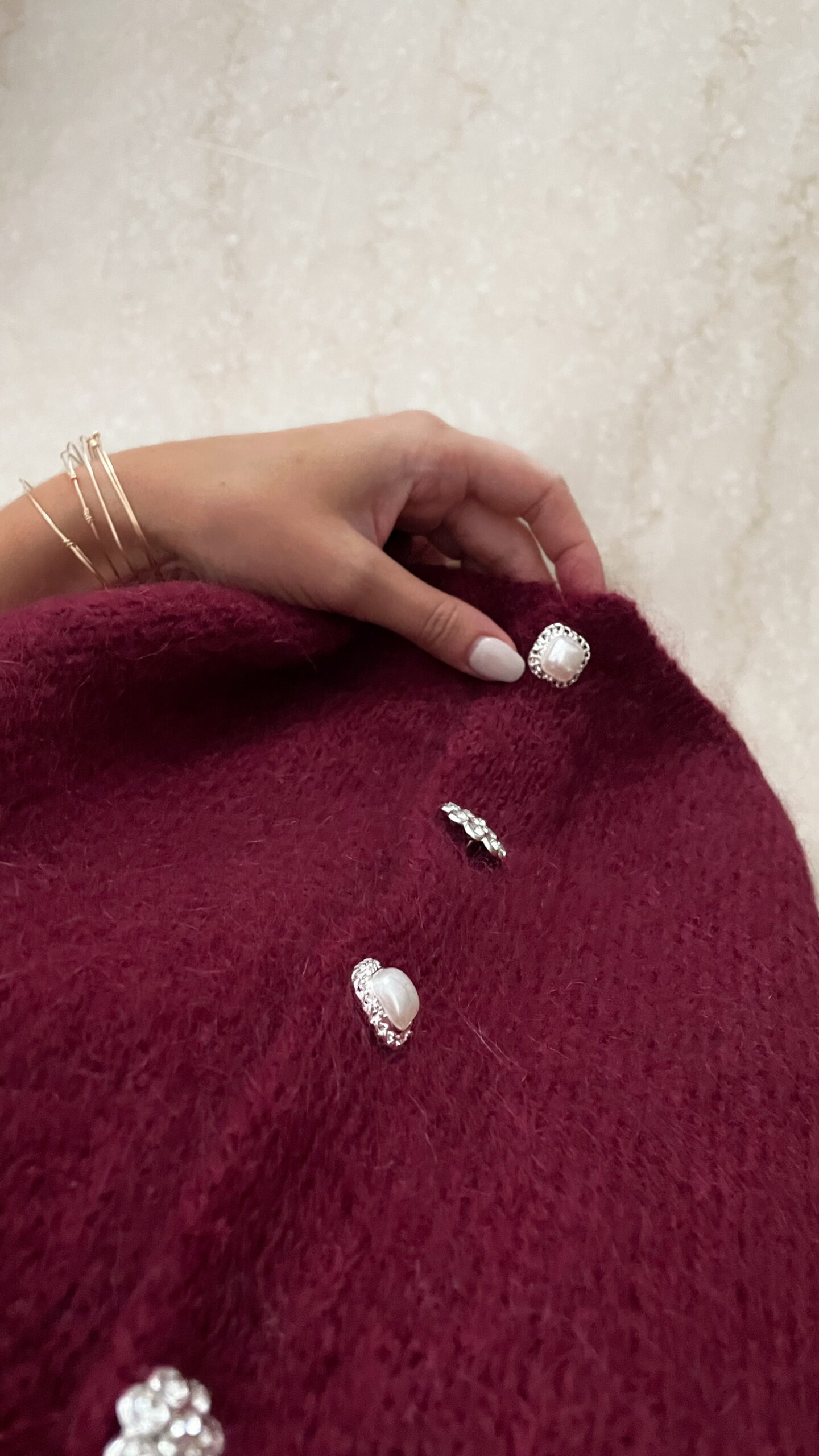 Femme portant un pull en mohair bordeaux à manches courtes, orné de boutons strass, associé à un foulard beige à motifs et des bijoux dorés au poignet.