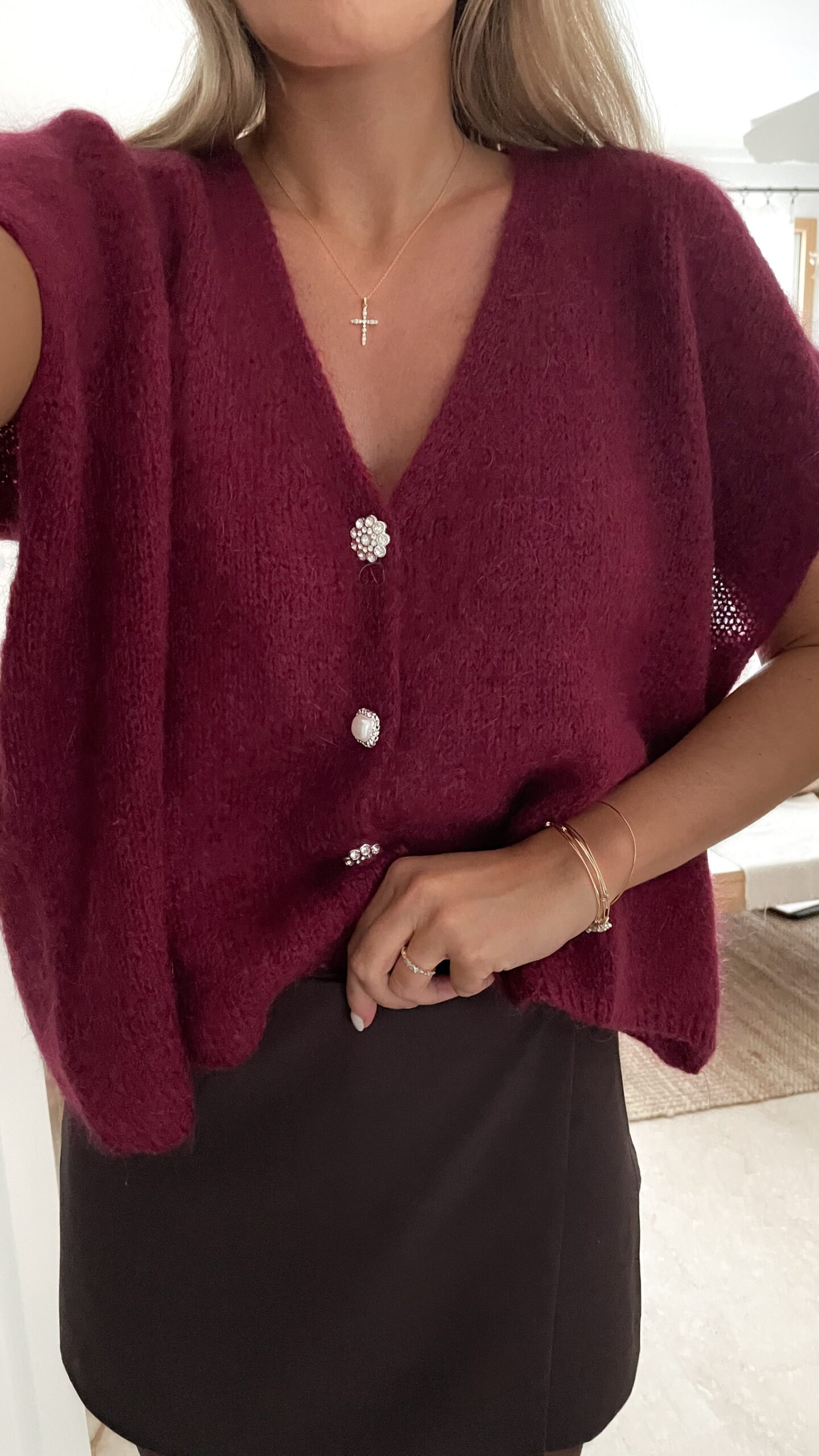 Femme portant un pull en mohair bordeaux à manches courtes, orné de boutons strass, associé à un foulard beige à motifs et des bijoux dorés au poignet.