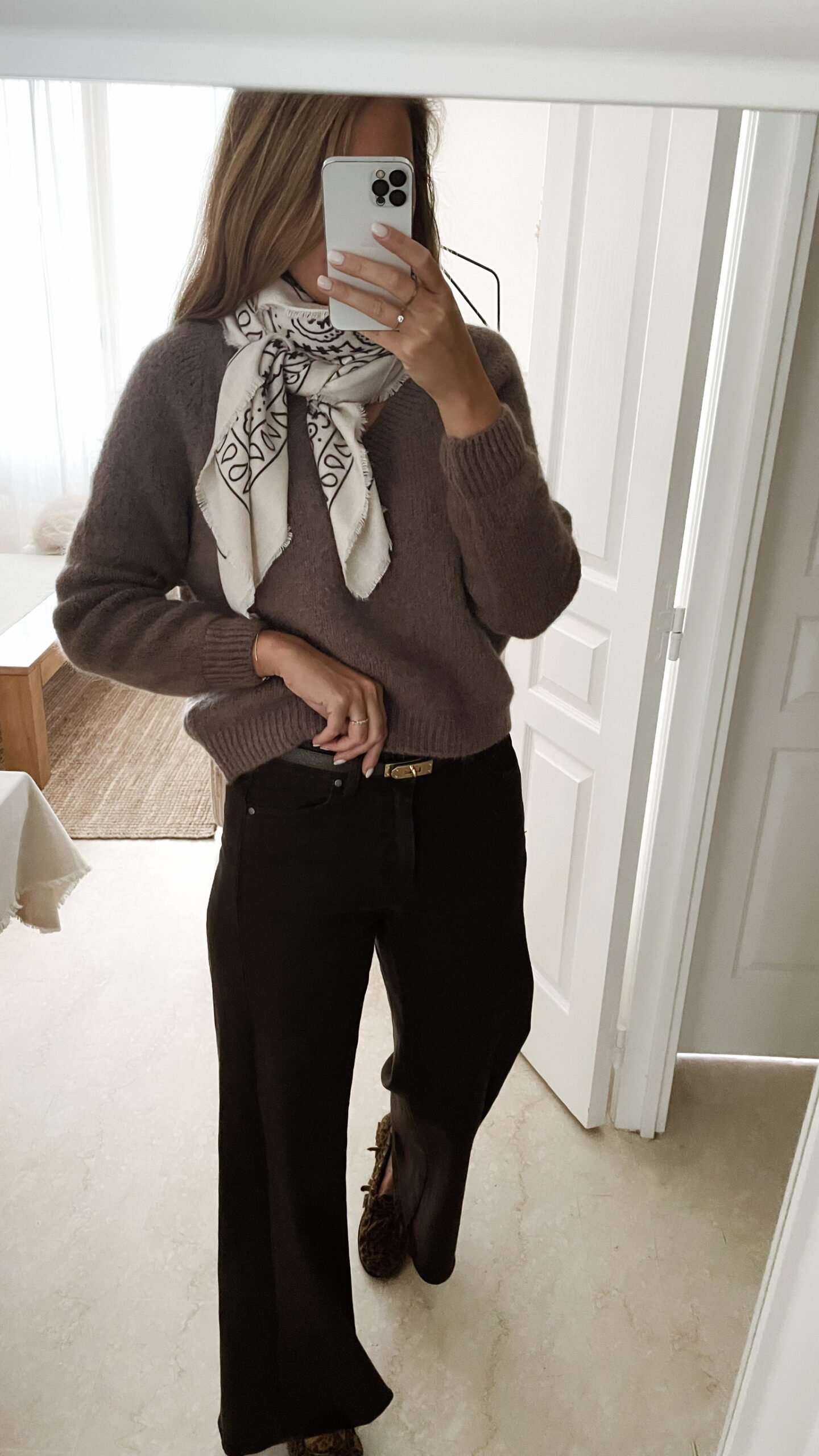 Femme portant un pull doux en alpaga couleur taupe, associé à un pantalon foncé à jambe large, une ceinture fine dorée, un foulard beige à motifs et des mocassins imprimés léopard.