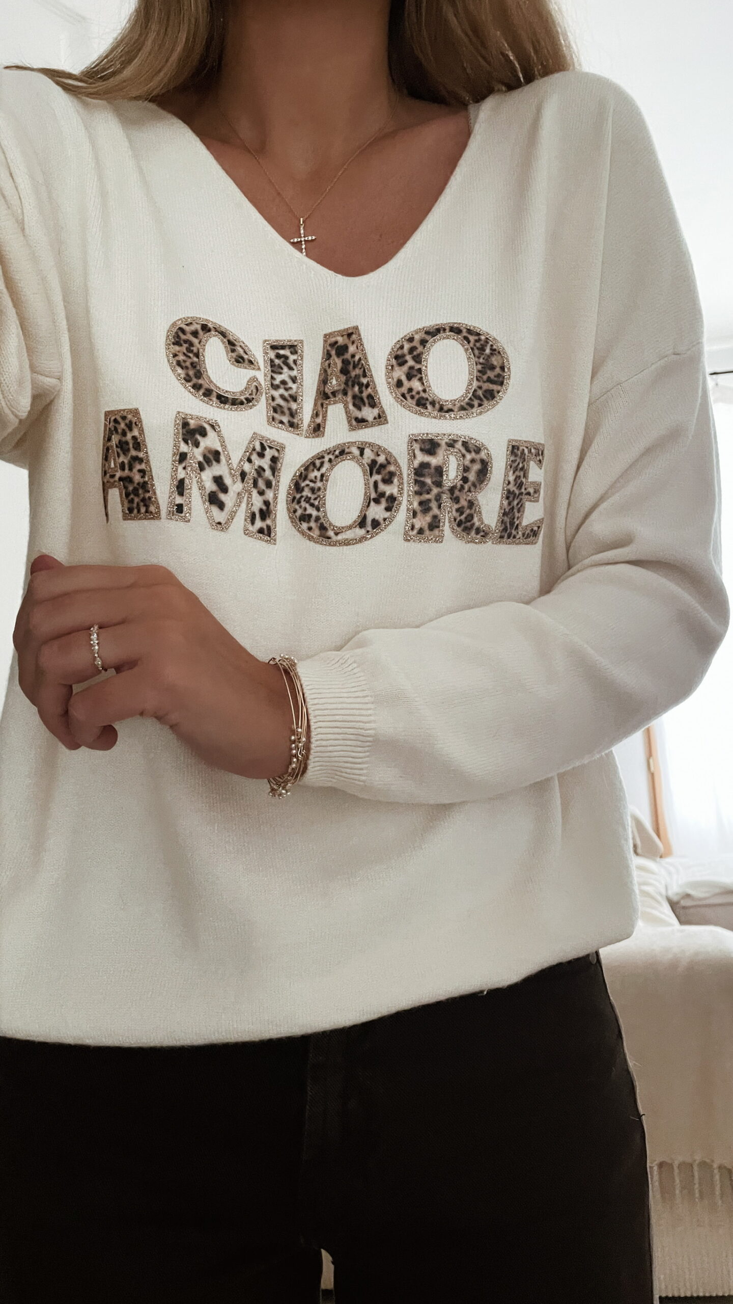 Femme portant un pull écru “Ciao Amore” avec un pantalon foncé, une veste en daim taupe et un sac à franges, pour un look automnal élégant et décontracté.