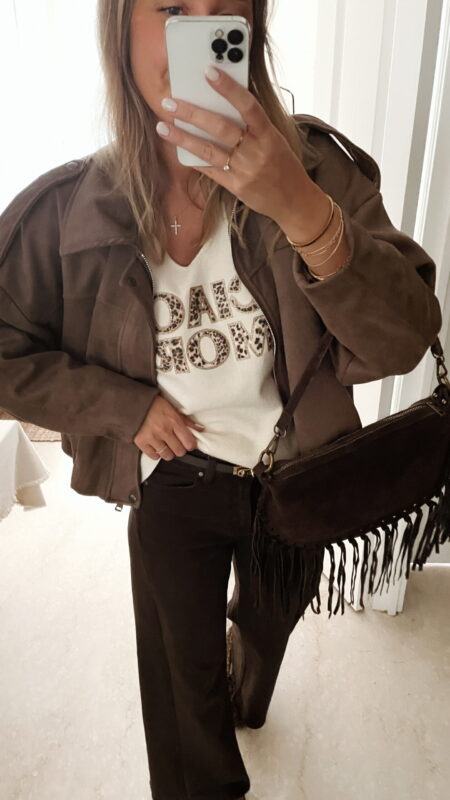 Femme portant une veste effet daim couleur marron taupe, assortie à un pull écru avec inscription "ciao amore", un pantalon marron, et un sac en daim marron avec des franges dans un intérieur lumineux.