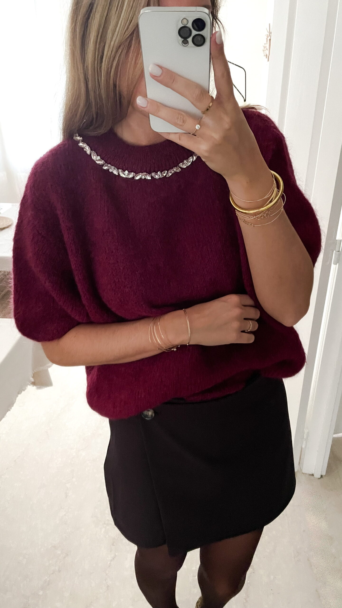 Femme portant un pull en maille bordeaux orné de strass au col, associé à une jupe noire courte et des bijoux dorés, photographiée devant un miroir.