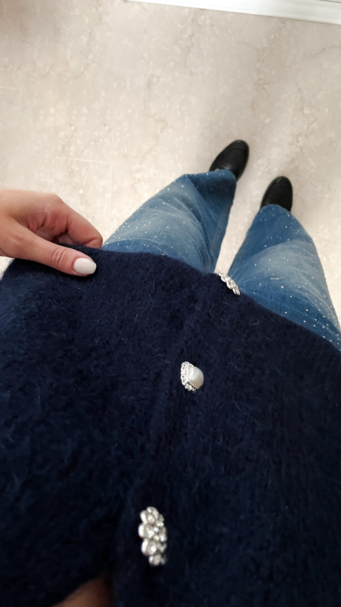 Femme portant un pull bleu en mohair à boutons bijoux et un pantalon en jean bleu foncé orné de strass, photographiée dans un intérieur lumineux.