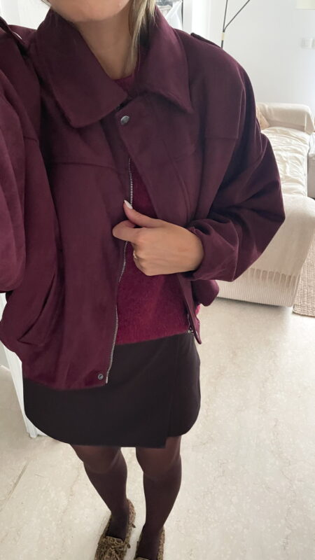 Femme portant une veste effet daim couleur bordeaux, assortie à un pull ton sur ton, une jupe noire et des collants foncés, dans un intérieur lumineux.
