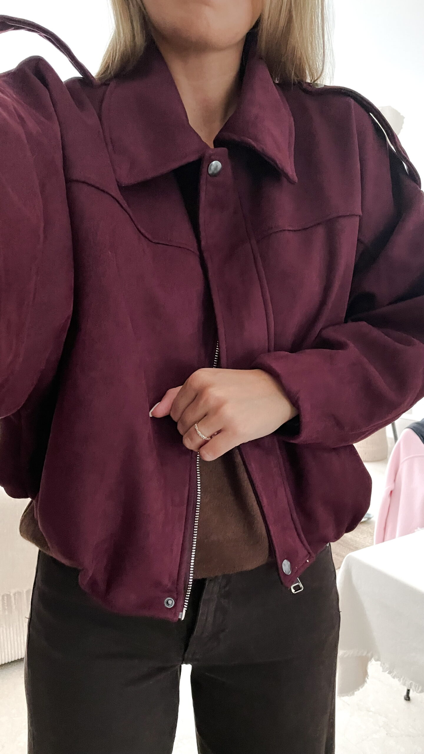 Femme portant une veste effet daim couleur bordeaux, assortie à un pull ton sur ton, une jupe noire et des collants foncés, dans un intérieur lumineux.