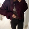 Femme portant une veste effet daim couleur bordeaux, assortie à un pull ton sur ton, une jupe noire et des collants foncés, dans un intérieur lumineux.