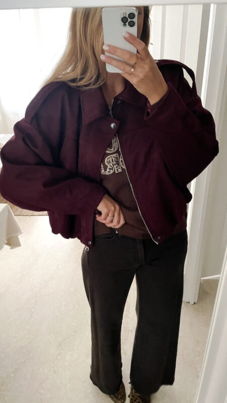 Femme portant une veste effet daim couleur bordeaux, assortie à un pull ton sur ton, une jupe noire et des collants foncés, dans un intérieur lumineux.